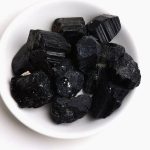 Vários cristais de turmalina negra em bruto, com formas irregulares e superfícies estriadas, apresentados numa tigela branca.