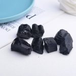 Vários fragmentos de turmalina negra bruta com superfícies irregulares e brilho natural, colocados sobre uma superfície branca ao lado de uma tigela azul.