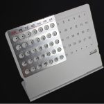 Calendário perpétuo de metal com dias, meses e semanas, exibido numa base de suporte branco, com detalhes em cinza e letras em vermelho e preto.