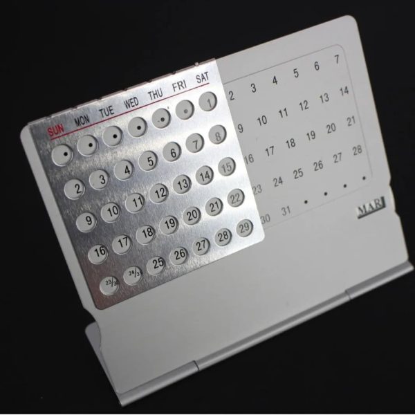 Calendário perpétuo de metal com dias, meses e semanas, exibido numa base de suporte branco, com detalhes em cinza e letras em vermelho e preto.