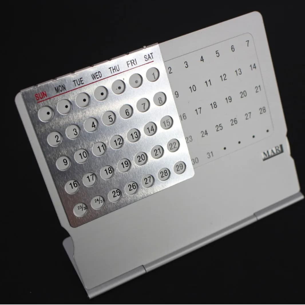 Calendário Perpétuo em Metal Calendário perpétuo de metal com dias, meses e semanas, exibido numa base de suporte branco, com detalhes em cinza e letras em vermelho e preto.