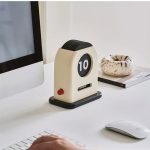 Relógio de mesa com display digital de número grande, base preta e corpo branco, colocado numa secretária com computador, livro e objeto decorativo.