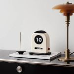 Relógio de mesa com visor digital mostrando o número 10, ao lado de uma lâmpada de mesa dourada e uma vela com incenso, sobre uma pilha de livros.
