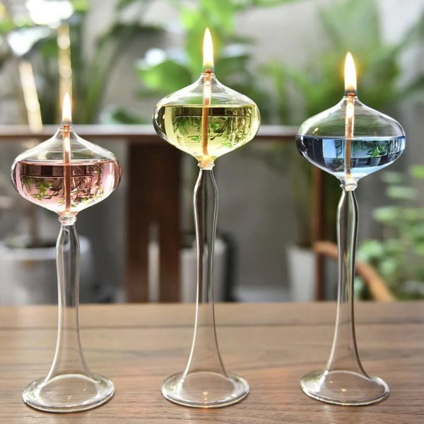 Três candeeiros de óleo de vidro com diferentes cores de líquido, acesos, em linha numa mesa de madeira com fundo de plantas verdes.