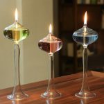Três candeeiros de mesa a óleo com diferentes cores de líquidos, cada um com uma chama acesa, colocados numa superfície de madeira.