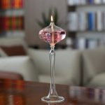 Candeeiro de mesa a óleo com base de vidro transparente, chama acesa, em ambiente de sala com livros e plantas ao fundo.