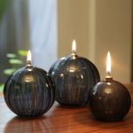 Três candeeiros de mesa a óleo em forma de bola com chama acesa, feitos de vidro escuro, colocados numa superfície de madeira, com fundo desfocado.