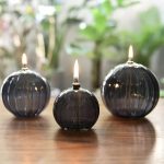 Três candeeiros de mesa a óleo em forma de bola, com chama acesa, colocados sobre uma superfície de madeira, com fundo de plantas verdes difusas.