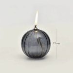 Candeeiro de mesa em forma de bola de vidro escuro com chama de óleo acesa, altura de 12cm, com detalhes de metal na parte superior.