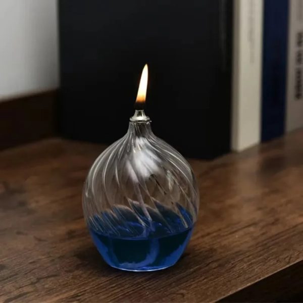 Candeeiro de mesa a óleo com design ondulado de vidro transparente, com líquido azul na base, aceso, colocado numa superfície de madeira junto a livros.