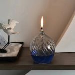 Candeeiro de mesa a óleo com design moderno, vidro transparente com detalhes ondulados, com chama acesa, colocado numa superfície de madeira.