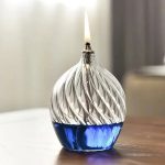 Candeeiro de mesa a óleo com design ondulado de vidro transparente, com chama artificial acesa na parte superior, colocado numa superfície de madeira.