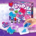 Conjunto de ingredientes e moldes para fazer sabão colorido e com glitter, incluindo líquidos, moldes de borboleta e coração, e uma mão a despejar o líquido azul.