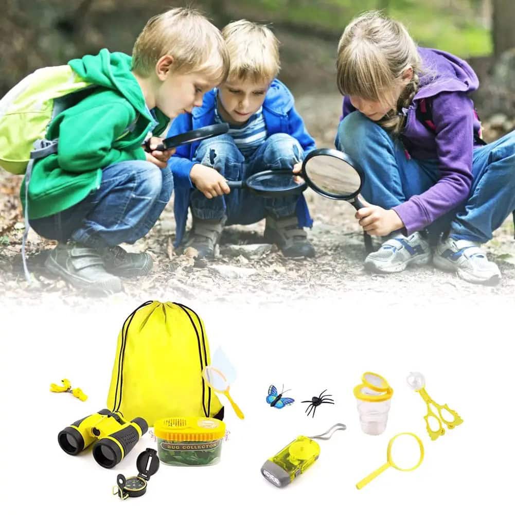 Três crianças explorando ao ar livre com lupa, observando o solo e pequenos objetos, com um kit de exploração científico ao lado.