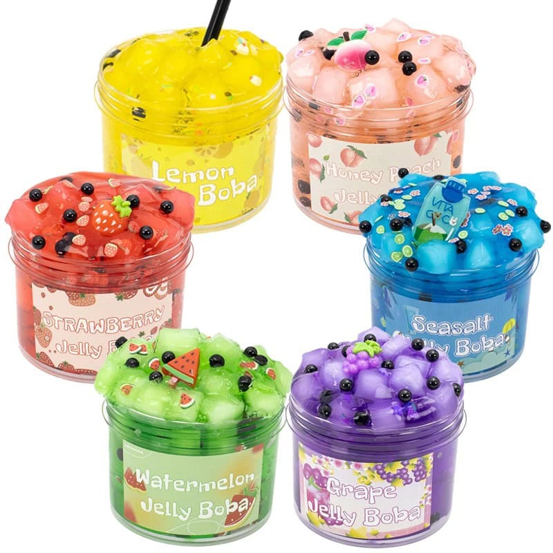 Vários potes de slime colorido com diferentes aromas e decorativos, incluindo frutas de plástico, glitter e pérolas, em frascos transparentes com etiquetas temáticas.