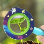 Lupa infantil com moldura decorada com estrelas e astronauta, focada numa folha verde jovem, mostrando detalhes da textura.
