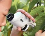 Jovem a observar folhas com microscópio portátil, focando na amostra, com fundo de plantas verdes.