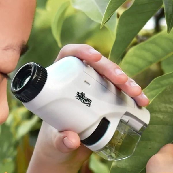 Jovem a observar folhas com microscópio portátil, focando na amostra, com fundo de plantas verdes.