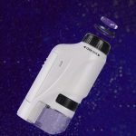 Microscópio portátil com lente de aumento ajustável, corpo branco, base transparente e lente superior destacável, sobre fundo estrelado.