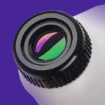 Lente de câmera com revestimento antirreflexo refletindo cores verdes e roxas, vista de cima com fundo roxo e branco.