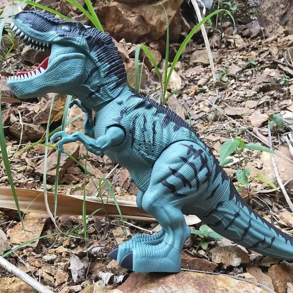 Dinossauro T-Rex de brinquedo com escamas azuis e detalhes em preto, posicionado em ambiente natural com terra, pedras e plantas ao fundo.