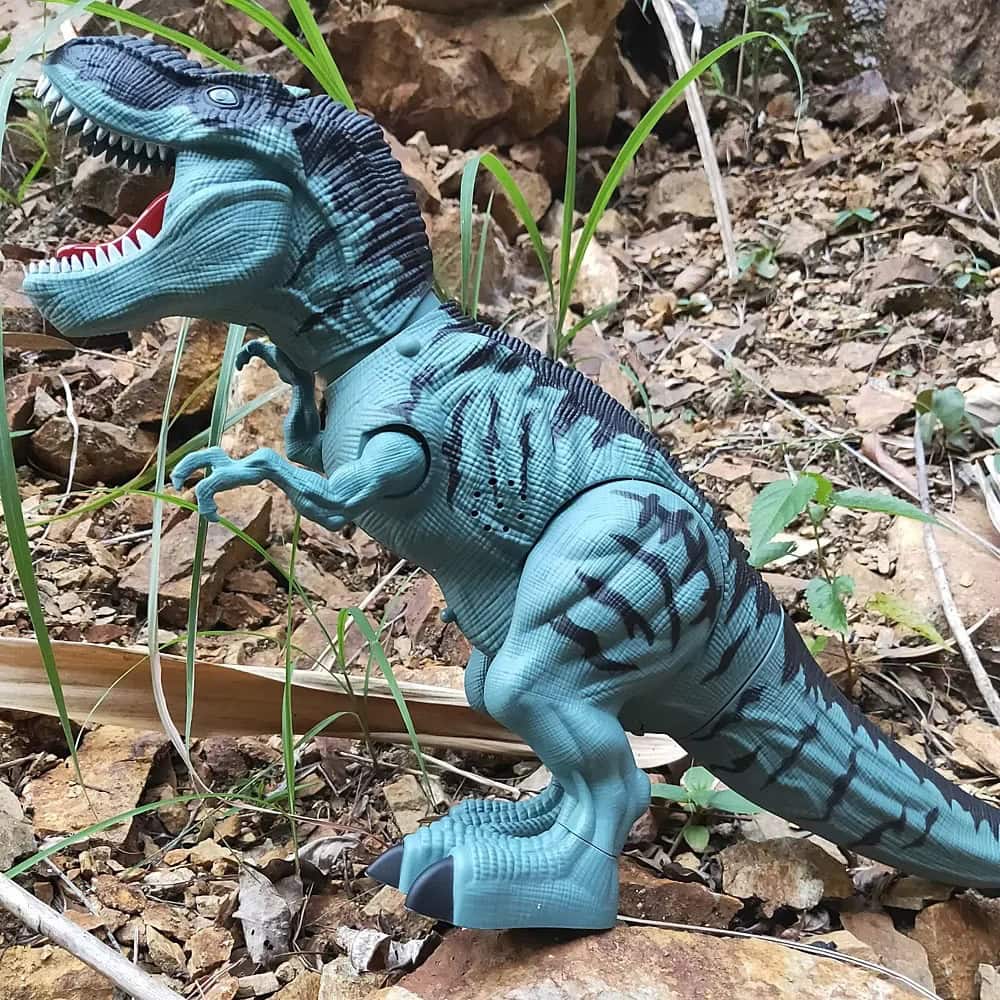 Dinossauro T-Rex de brinquedo ao ar livre Dinossauro T-Rex de brinquedo com escamas azuis e detalhes em preto, posicionado em ambiente natural com terra, pedras e plantas ao fundo.