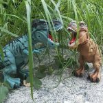 Dois dinossauros de brinquedo, um verde e outro castanho, com bocas abertas, rodeados por vegetação alta e folhas no chão.