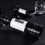 Telescópio com corpo branco e lentes pretas, colocado numa superfície escura com gotas de água, acompanhado de um copo de água transparente ao lado.