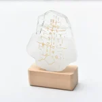 Escultura de vidro transparente com inscrições douradas, apoiada numa base de madeira, com fundo branco.