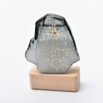 Pequena escultura de mineral com inscrições douradas, colocada sobre base de madeira clara, vista frontal com detalhes das inscrições.