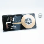 Placa de circuito com componentes eletrónicos, uma esfera metálica e uma placa circular dourada com logótipo de ciência, sobre fundo branco.