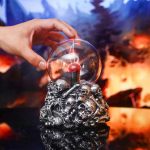 Bola de plasma com design de caveira, refletindo luzes coloridas, apoiada numa base decorativa com crânios de metal, ambiente com fundo de fogo e fumaça.