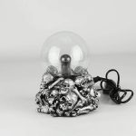 Bola de plasma com base de caveira em metal prateado, esfera de vidro transparente com descarga elétrica no interior, ligada a cabo elétrico preto.