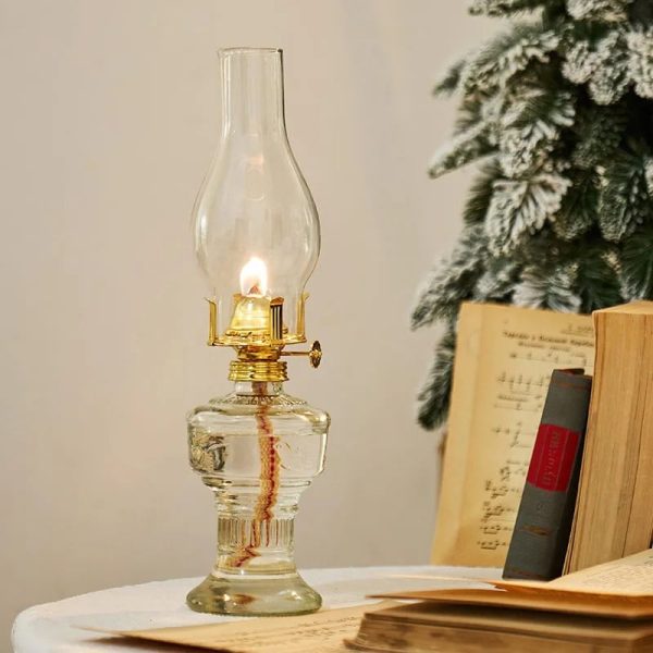 Candeeiro a óleo antigo de vidro transparente com chama acesa, ao lado de livros abertos e uma árvore de Natal decorada com neve artificial.