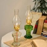 Dois candeeiros a óleo antigos com vidro transparente e base de vidro verde, acesos, colocados numa mesa junto a livros e uma árvore de Natal decorada.
