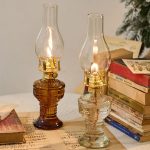 Dois candeeiros a óleo antigos com vidro transparente e chama acesa, colocados sobre uma mesa com livros antigos ao fundo.