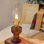 Candeeiro a óleo antigo com chama acesa, base de vidro âmbar e tubo de vidro transparente, em cima de livros antigos e documentos.