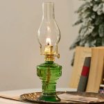 Candeeiro a óleo antigo com base de vidro verde e chama acesa, em ambiente interior com livros e árvore decorativa ao fundo.