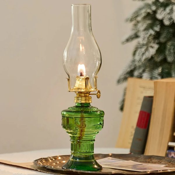 Candeeiro a óleo antigo com base de vidro verde e chama acesa, em ambiente interior com livros e árvore decorativa ao fundo.