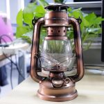 Candeeiro a óleo de estilo vintage com acabamento em bronze, com vidro transparente e alça de metal, colocado numa mesa de escritório com plantas ao fundo.