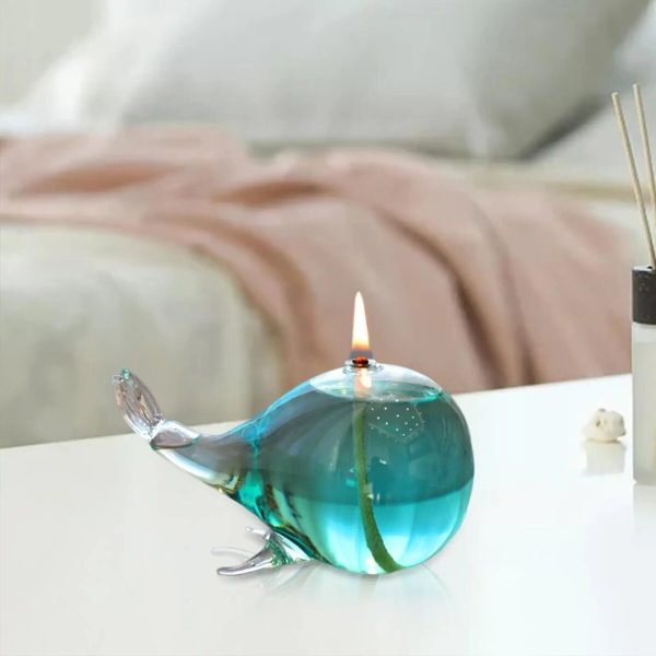 Luminária de óleo em forma de baleia de vidro com chama acesa, colocada numa mesa branca com fundo desfocado de cama e roupa de cama.