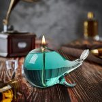 Candeeiro a óleo em forma de baleia de vidro com chama acesa, colocado numa mesa de madeira com fundo de parede cinzenta.