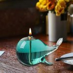 Candeeiro a óleo em forma de baleia de vidro com chama acesa, colocado numa mesa de madeira, com flores ao fundo.