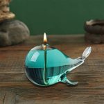 Candeeiro a óleo em forma de baleia de vidro azul com vela acesa, colocado numa superfície de madeira com fundo verde.