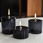 Três velas de vidro negras acesas em diferentes tamanhos, colocadas numa superfície branca, com fundo escuro e ambiente acolhedor.