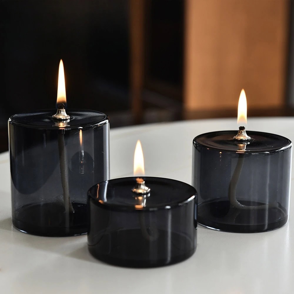Velas de vidro negras acesas Três velas de vidro negras acesas em diferentes tamanhos, colocadas numa superfície branca, com fundo escuro e ambiente acolhedor.
