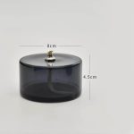 Candeeiro a óleo cilíndrico de vidro escuro com pequena chama acesa no topo, medindo 8cm de diâmetro e 4.5cm de altura.