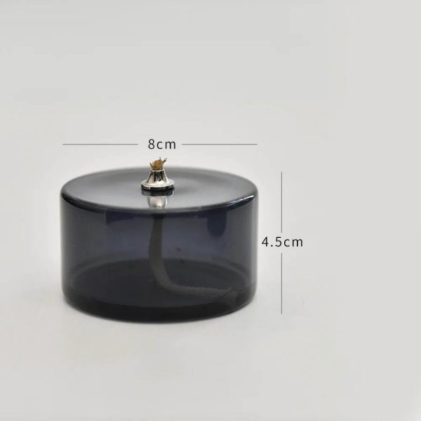 Candeeiro a óleo cilíndrico de vidro escuro com pequena chama acesa no topo, medindo 8cm de diâmetro e 4.5cm de altura.