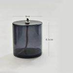 Candeeiro a óleo cilíndrico de vidro escuro com tampa metálica e tubo de ignição visível, dimensões indicadas de 8cm de diâmetro e 8,5cm de altura.