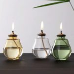 Três velas acesas em frascos de vidro de diferentes cores: amarelo, transparente e verde, com pavios visíveis, sobre uma superfície escura com fundo branco.